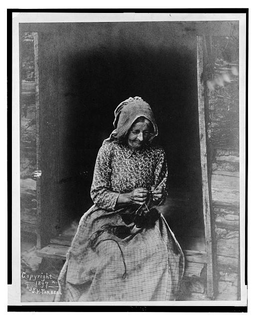 An 1897 photograph of an elderly woman in a bonnet and print dress, knitting in the doorway of a log cabin