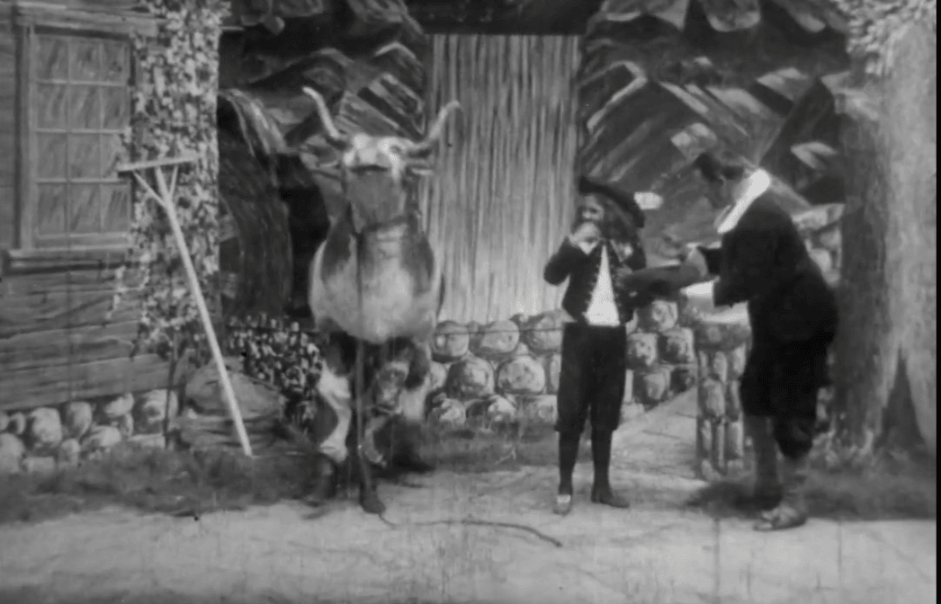 Black-and-white film still on a painted stage set: a horned pantomime cow stands by a cottage while Jack, in cap and knee breeches, faces a cloaked man in a white ruff who holds out his hat
